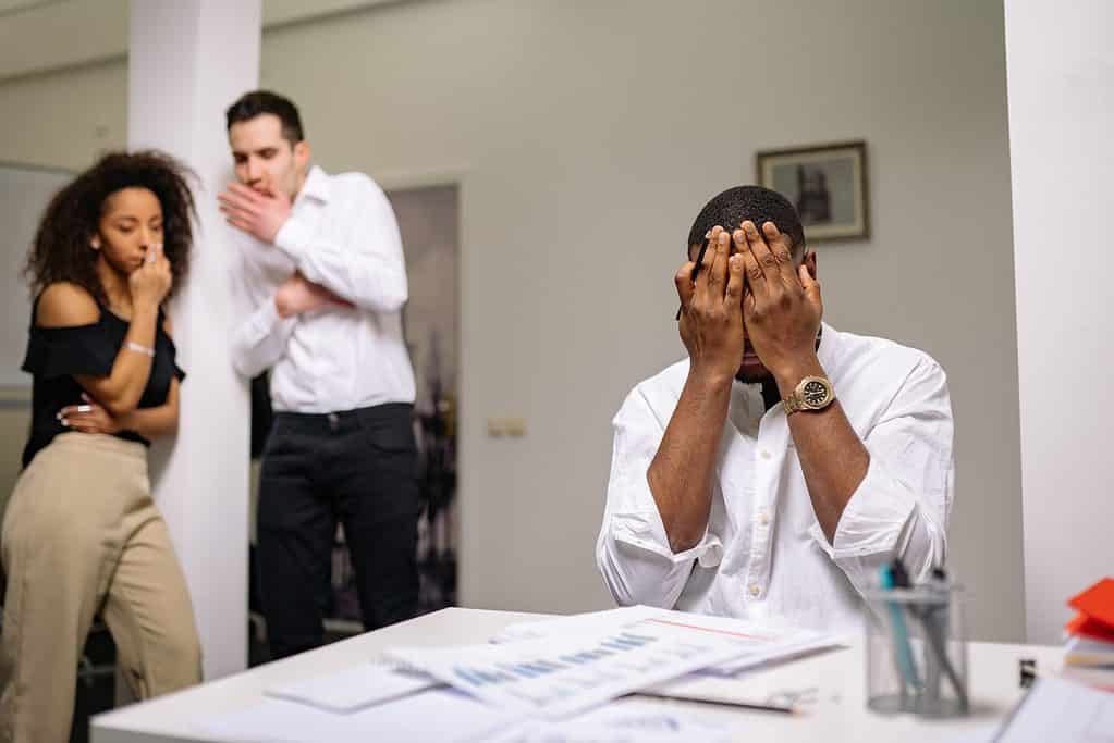 Drei Personen in einem Büro, Mann schlägt die Hände vor dem Gesicht zusammen, zwei Personen blicken sorgenvoll zu dem Mann–